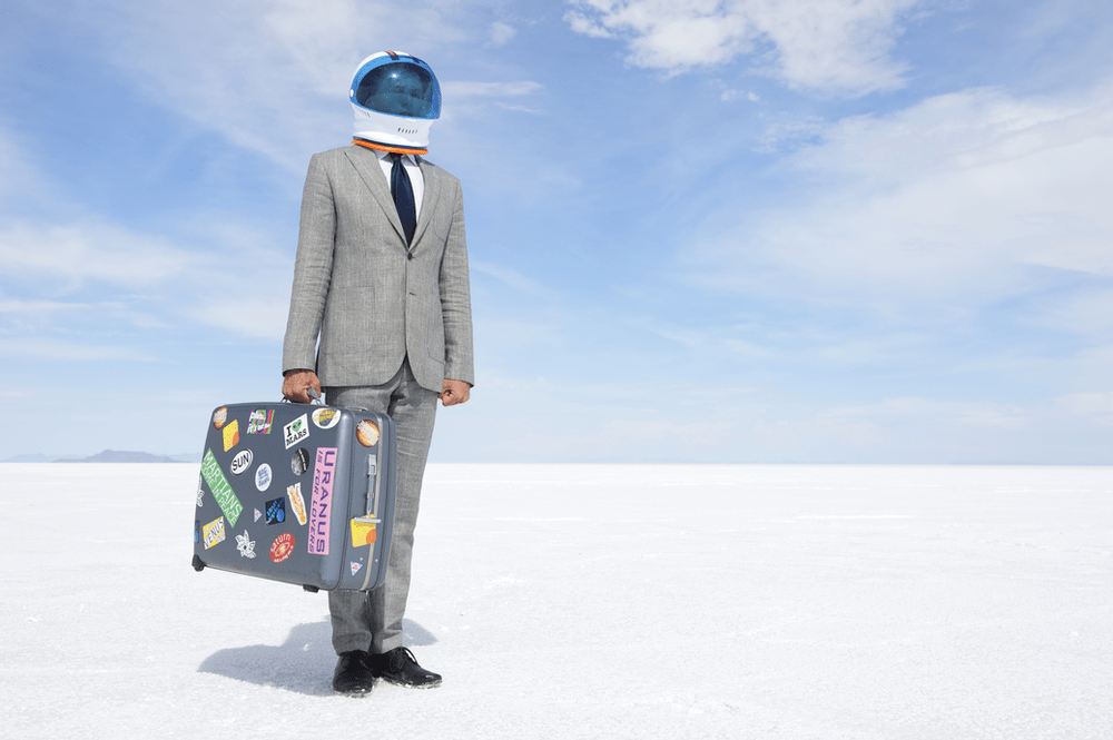 a man in a grey suit and an astronaut helmet holds a luggage with travel stickers; science fiction job