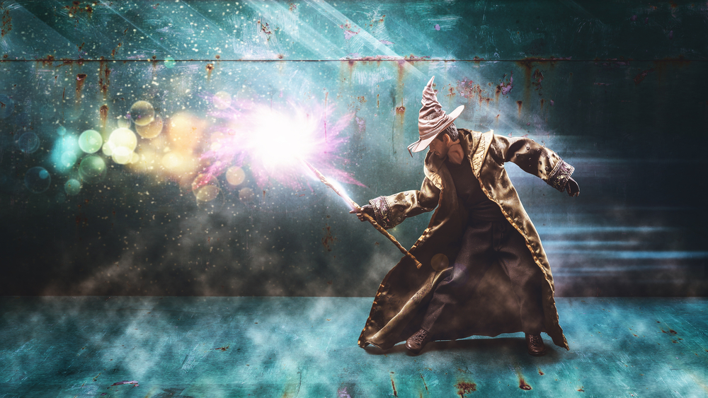 a man in a brown wizard hat, robes, and boots holds out a staff with a purple burst of magic coming out of it; fantasy job