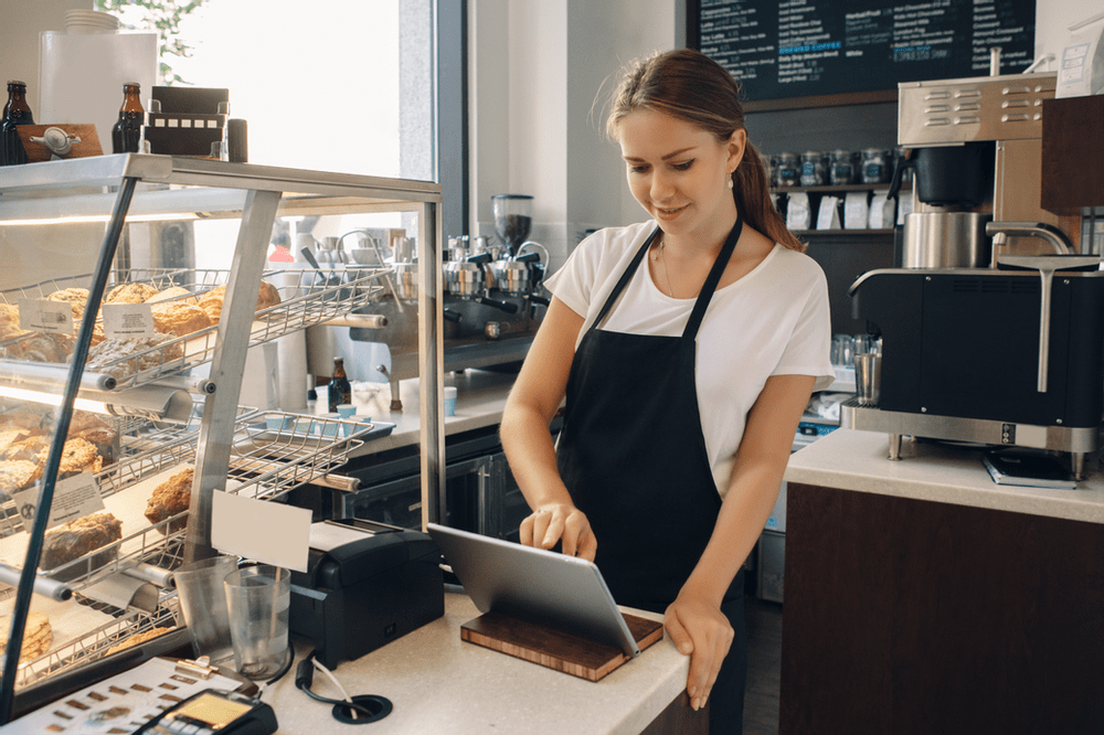 a young female barista taking orders on a tablet next to a case of baked goods and in front of a coffee machine and coffee menu