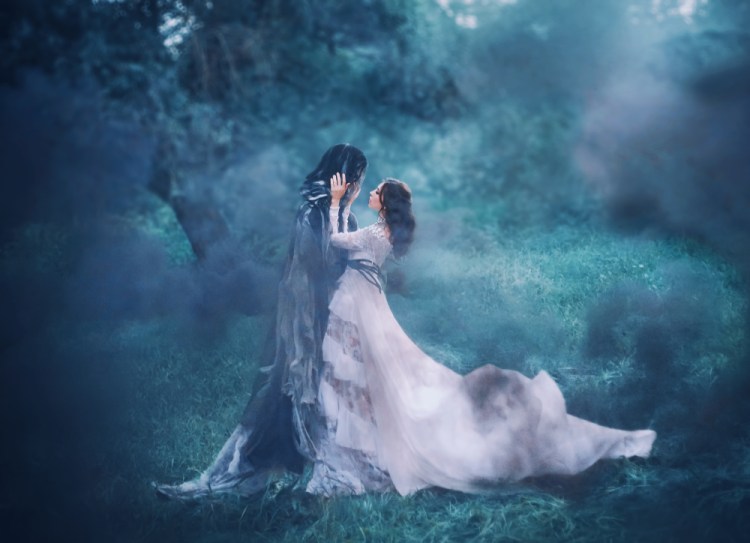 brunette woman in a white dress holding the face of a hooded figure