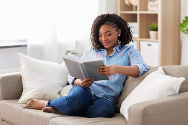 a Black woman sitting on a tan couch with white pillows reads a book; a wooden shookshelf behind her