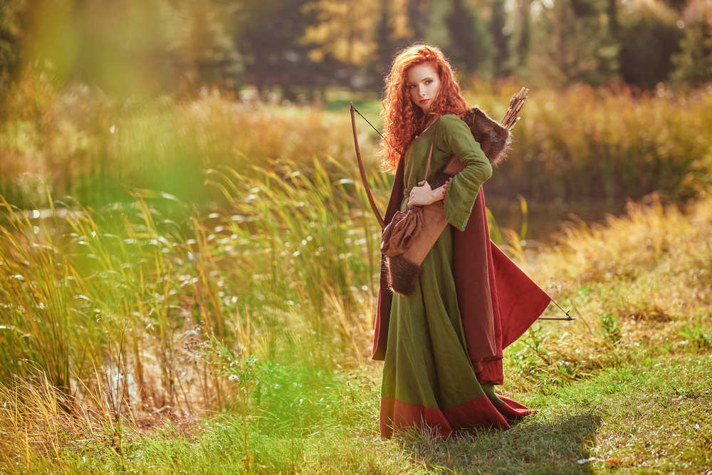 young woman archer with magnificent long red hair in a historical celtic dress is posing near the lake